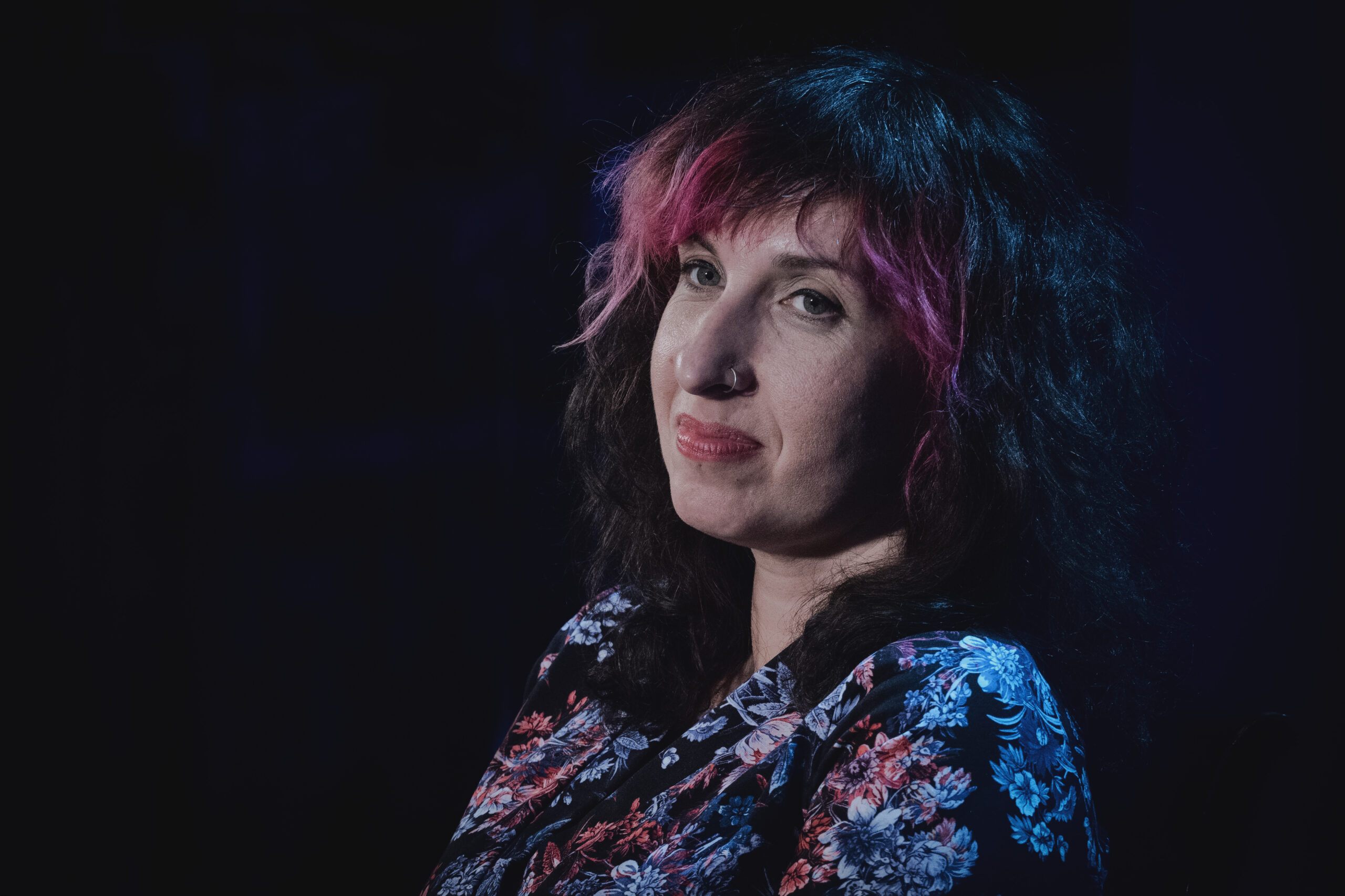 Zdjęcie portretowe ciemnowłosej kobiety z różową grzywką. Jest ubrana w ciemną bluzkę w kolorowe kwiaty. Tło jest czarne.