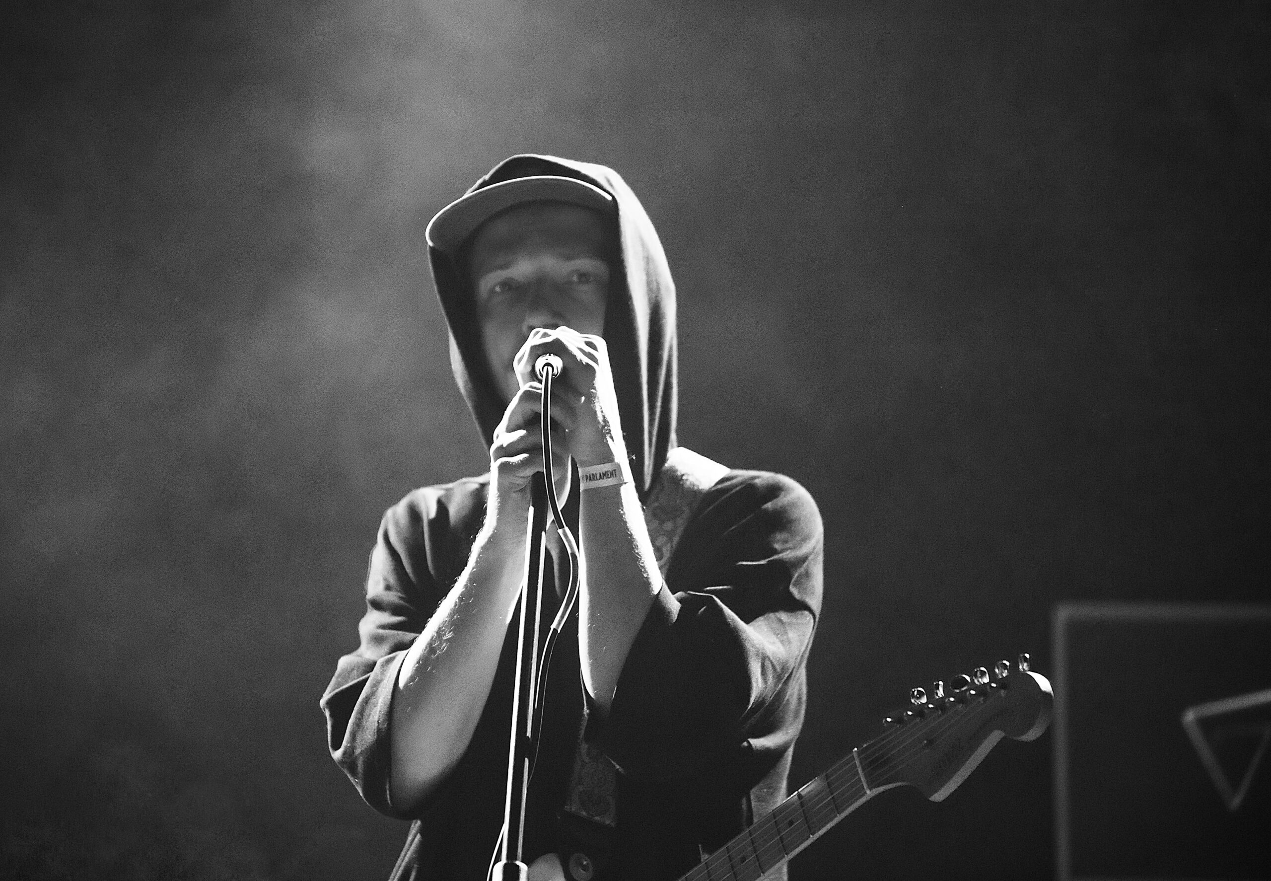 Czarno-białe zdjęcie młodego mężczyzny z mikrofonem na statywie i gitarą. Jest w ujęciu od pasa w górę. Obie dłonie trzyma na mikrofonie zbliżonym do ust. Przez ramię ma przewieszony pasek, na którym jest gitara zawieszona na wysokości jego brzucha. Mężczyzna jest ubrany w ciemną bluzę z kapturem założonym na głowę. Spod kaptura wystaje daszek kaszkietówki. Tło ciemne z delikatnymi promieniami w lewym górnym rogu i snującym się podświetlonym dymem.