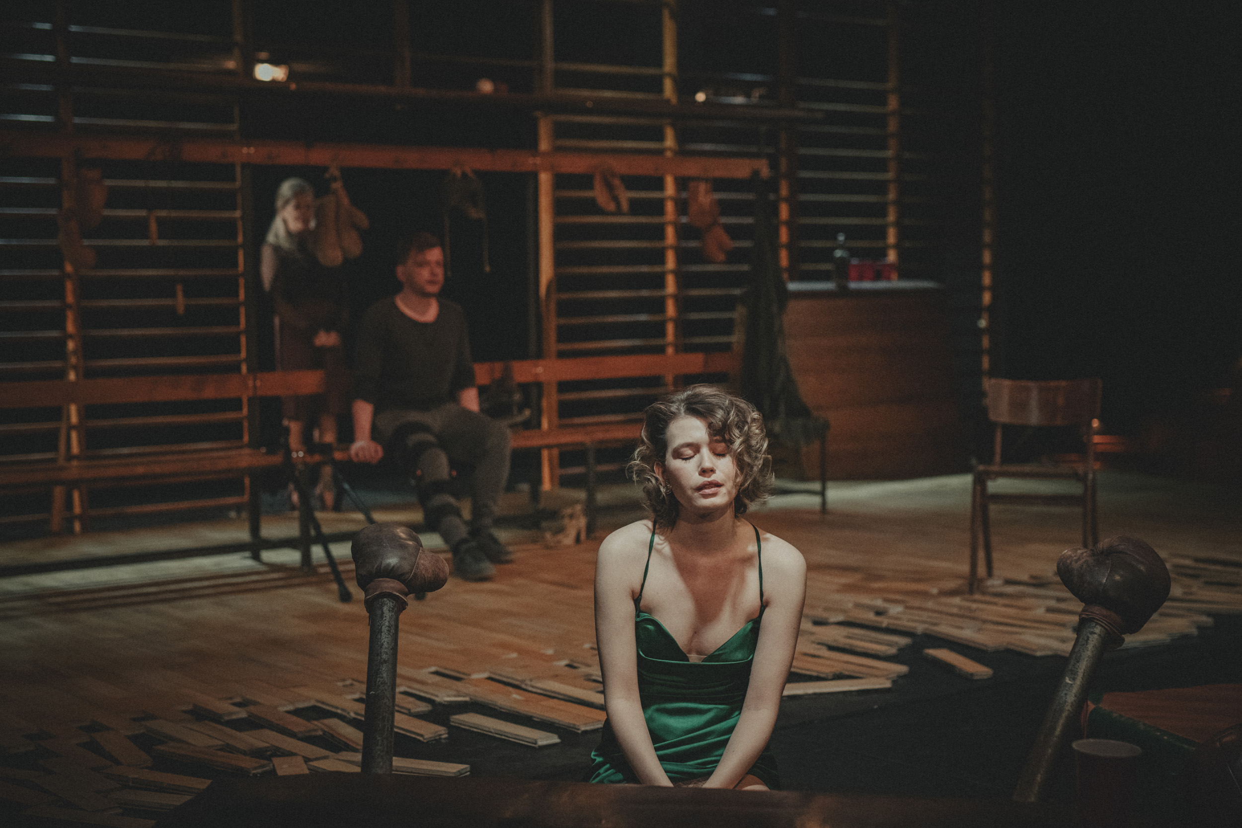 Zdjęcie trzech osób na scenie. Na pierwszym planie, wyraźnie oświetlona, młoda kobieta. Siedzi na scenie. Ma przymknięte oczy i rozchylone usta. Jest ubrana w ciemnozieloną, atłasową sukienkę. Ma ciemne, kręcone włosy sięgające za ucho. W rozmytym tle, w półmroku jeszcze dwie postacie. Mężczyzna siedzący na długiej drewnianej ławce. Tuż za nim stoi kobieta. Głównymi elementami scenografii są wysokie drewniane drabinki i parkiet z jasnych klepek, który rozpada się na brzegach.