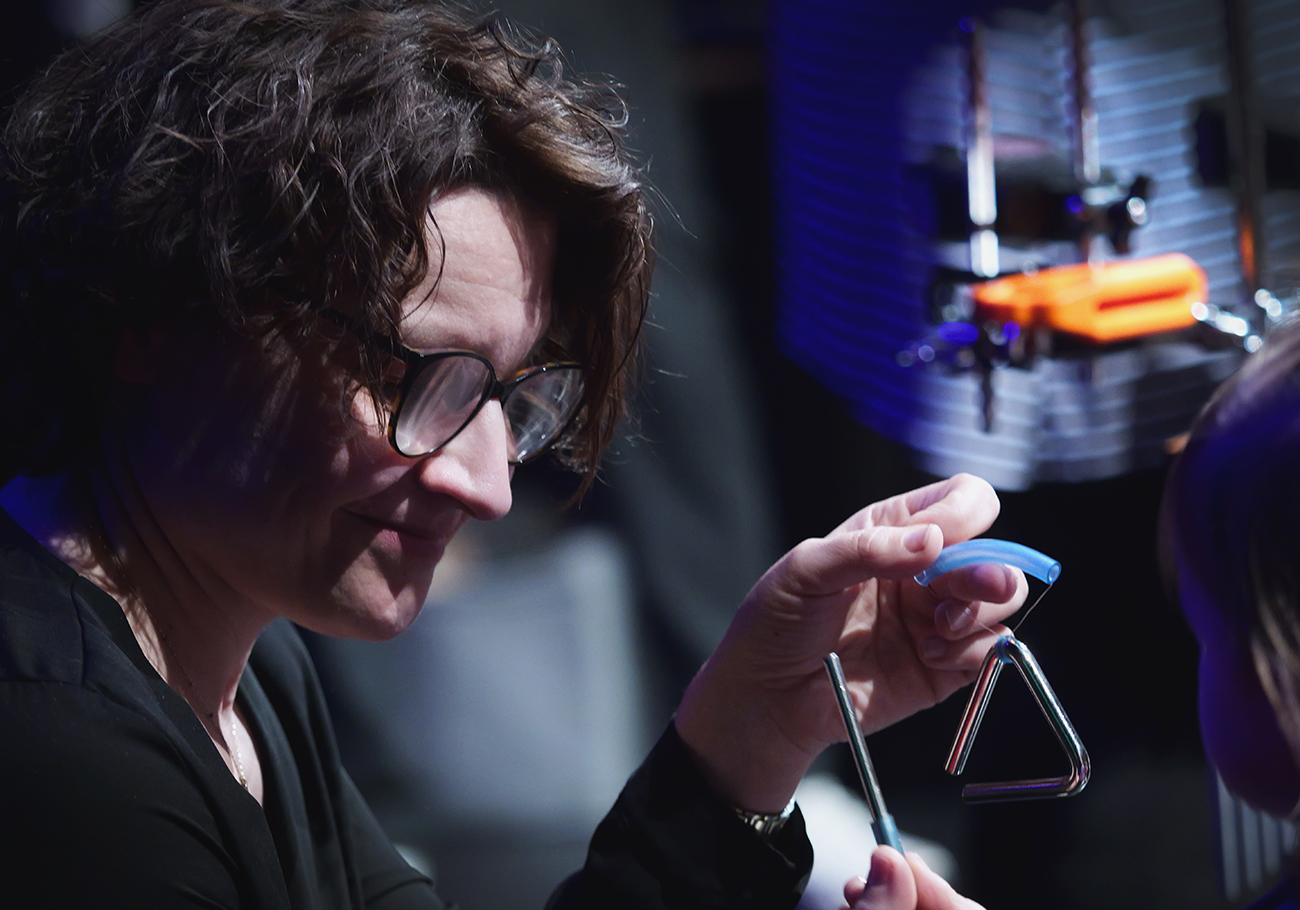 portret kobiety w średnim wieku w ujęciu z półprofilu. Ma kręcone włosy i okulary w prostokątnych, czarnych, grubych ramkach. W ręce trzyma dwa mały metalowy trójkąt (instrument) i pałeczkę.