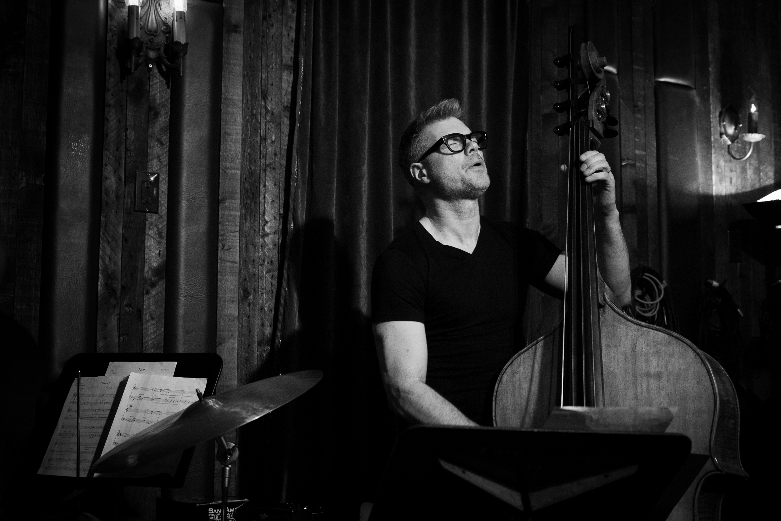 Czarno-białe zdjęcie mężczyzny grającego na wiolonczeli. Trzyma ją po prawej stronie, jedną rękę ma na gryfie, drugą niżej na strunach. Przed nim stoi pulpit na nuty. Mężczyzna ma krótkie włosy i okulary. Jest ubrany w czarną koszulkę z krótkimi rękawami. Po lewej stronie znajduje się talerz perkusyjny i jeszcze jeden pulpit z nutami. W tle ciemne pomieszczenie z kotarą. W prawym górnym rogu świecący kinkiet.