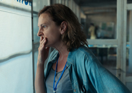 Zdjęcie kobiety w ujęciu z profilu. Kobieta jest ubrana w szarą koszulkę i zarzuconą na ramiona dżinsową koszulę. Na szyi ma zawieszoną niebieską smycz na identyfikator. Ma szeroko otwarte oczy, prawą dłoń przykłada do ust. Jest zwrócona w stronę okna zasłoniętego żaluzją. W rozmytym tle korytarz.