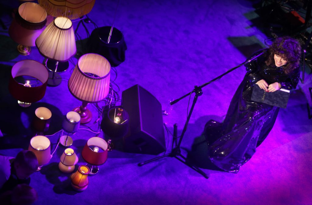 fot.W.Nieśpiałowski [Ujęcie z góry. Po lewej dużo lampek z abażurami w różnych kształtach i rozmiarach. Po prawej siedząca na krześle kobieta z kręconymi włosami, ubrana w długą czarną suknię, trzymająca przed sobą zamkniętą książkę. Tło fioletowe.]