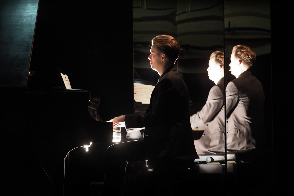 fot. Wojciech Nieśpiałowski / [Pianista w garniturze z dłońmi na klawiaturze fortepianu. W tle odbicia muzyka w trójdzielnym lustrze. W mroku punktowe światło pada tylko na lawiaturę, nuty i twarz mężczyzny.]