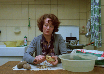 Zdjęcie kobiety siedzącej przy stole kuchennym. Kobieta obiera ziemniaki, na stole kuchennym leży ścierka na niej obierki. Nad obierkami dłonie kobiety trzymające częściowo obranego ziemniaka i nożyk. Po lewej na stole kilka nieobranych ziemniaków, po prawej półprzezroczysta, plastikowa miska z wodą. Kobieta jest ubrana w ciemnofioletową koszulę w jasne kwiaty i jasnoszary, rozpięty sweter, ma falowane, kasztanowe włosy sięgające za ucho. W tle ściana wyłożona beżowymi kafelkami. Pod ścianą po lewej biały zlew i żółta suszarka na naczynia. Po prawej kuchenka, na której stoją garnki z pokrywkami i patelnia.