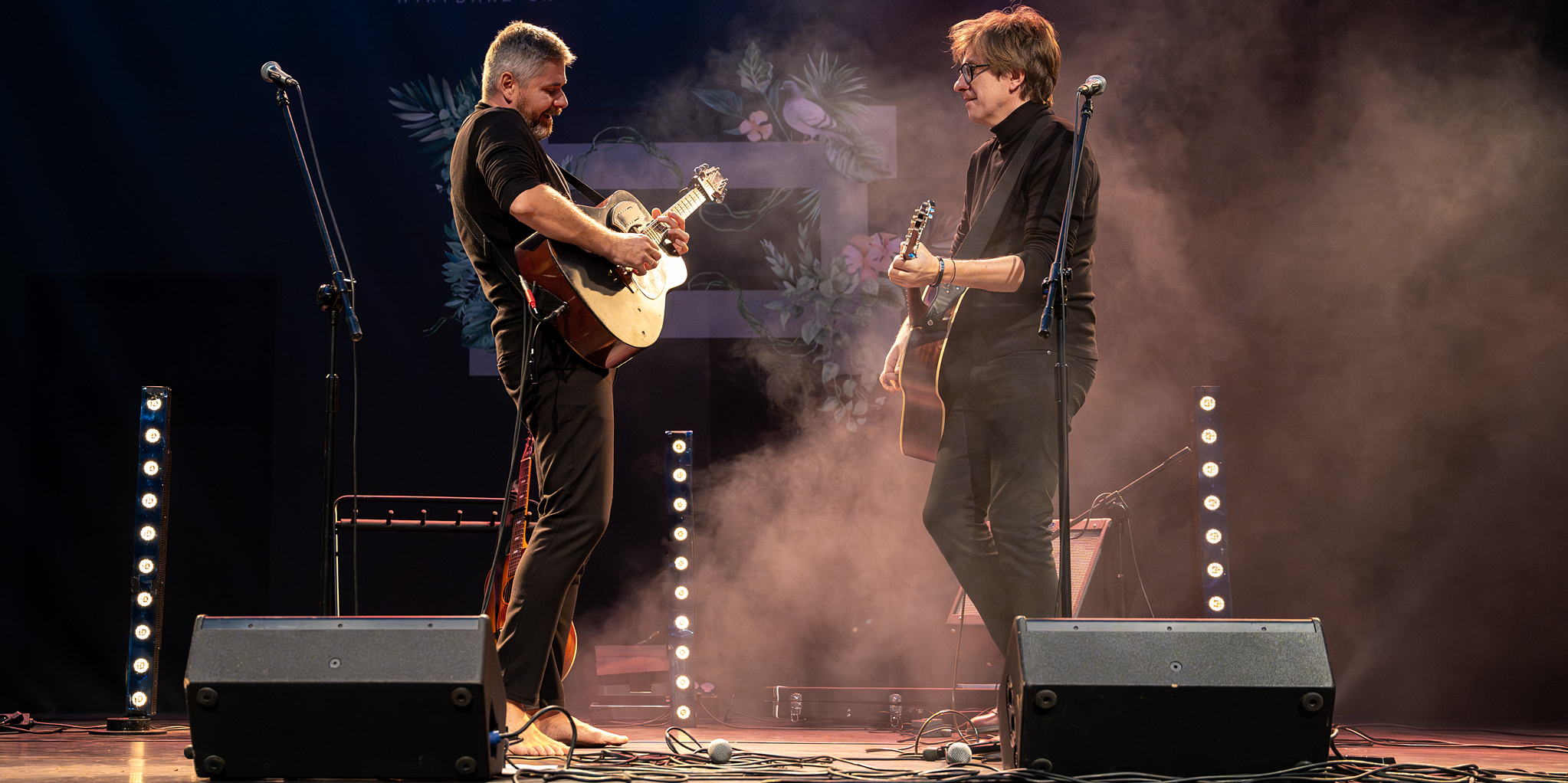 Zdjęcie dwóch mężczyzn w czarnych strojach. Stoją na scenie zwróceni do siebie nawzajem. Grają na gitarach. W przestrzeni unosi się sztuczny dym podświetlony światłem reflektorów. Tło ciemne.