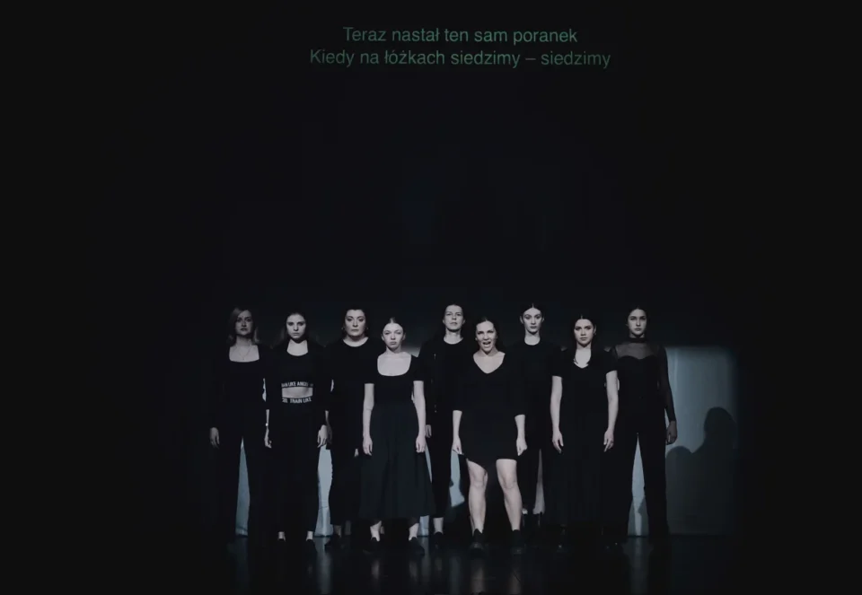 Grupa dziewięciu kobiet stojących na scenie. Wszystkie mają czarne stroje. Tło jest czarne.