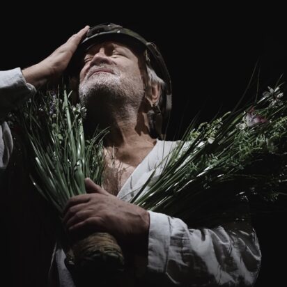 Obraz Główny: Zdjęcie Andrzeja Seweryna w roli króla Leara. Ma na sobie luźną sukmanę i kask na głowie. Jedną ręką przytula do klatki piersiowej dwa bukiety w stylu łąkowym. Drugą ręką dotyka głowy. Twarz zwraca ku górze, ma zamknięte oczy. Tło jest czarne.