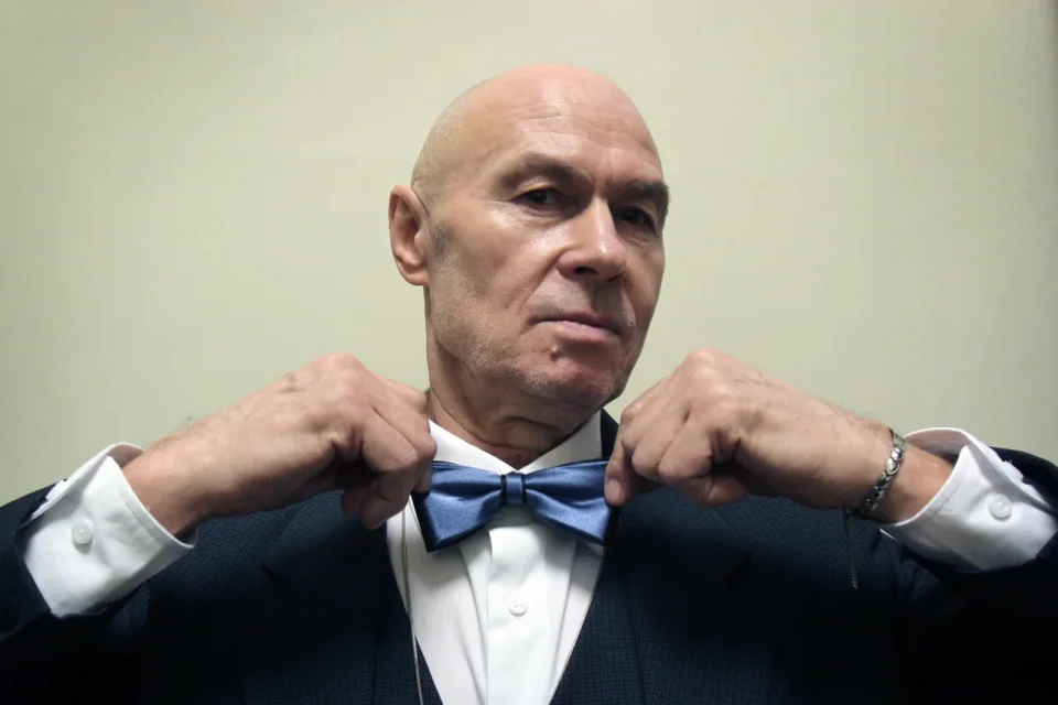 Zdjęcie portretowe łysego mężczyzny w garniturze z niebieską muchą. Przytrzymuje muchę palcami z obu stron. Tło jasnożółte.