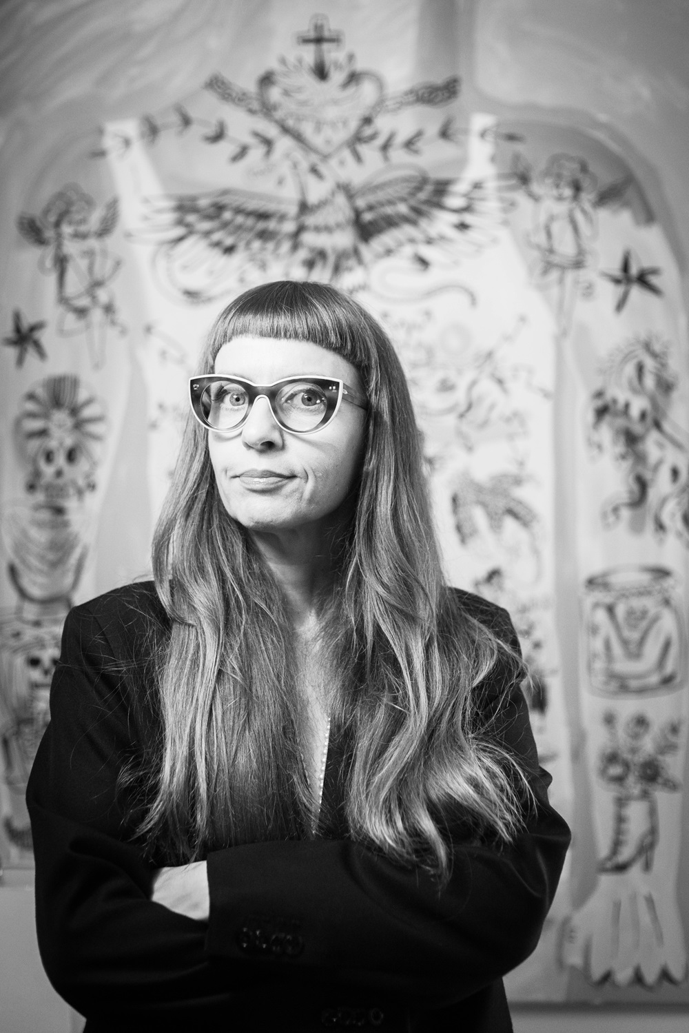 Czarno-białe zdjęcie kobiety w ujęciu portretowym. Kobieta jest ubrana w czarną marynarkę. Ma długie rozpuszczone włosy z krótką grzywką. Ma okulary z dużymi, grubymi oprawkami. W rozmytym tle jasna ściana w ciemne wzory.