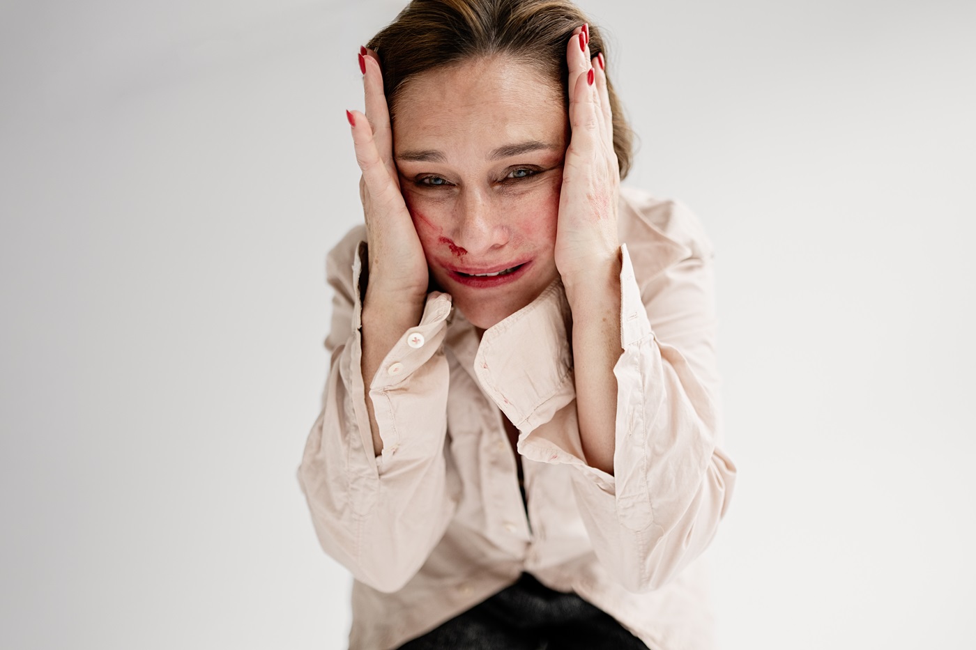 Zdjęcie kobiety z przestraszoną miną i plamką krwi w okolicach nosa. Kobieta przyciska dłonie do głowy po obu stronach twarzy. Jest ubrana w jasnobeżową, luźną koszulę z długimi rękawami i rozpiętymi mankietami. Tło jasnoszare.