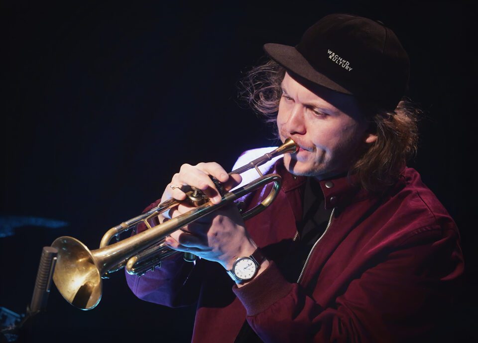 Zdjęcie mężczyzny grającego na trąbce. Jest ubrany w bordową bluzę ze stójką. Ma długie włosy wystające spod czarnej czapki z daszkiem. Tło ciemne.