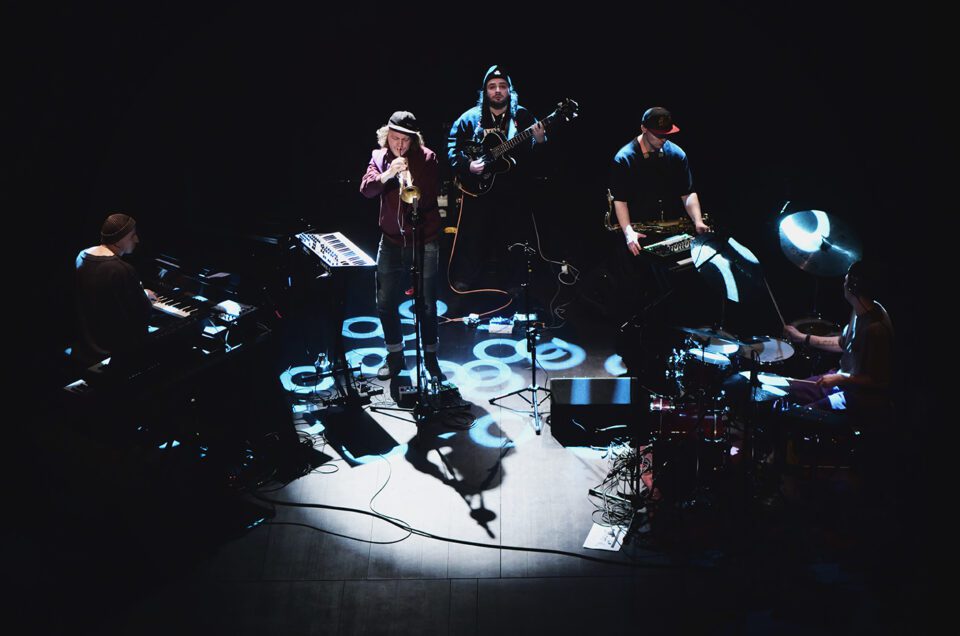 Zdjęcie pięcioosobowego zespołu na scenie. Klawiszowiec, trębacz, basista, saksofonista i perkusista. Na scenie refleksy białoniebieskiego światła. Tło ciemne.