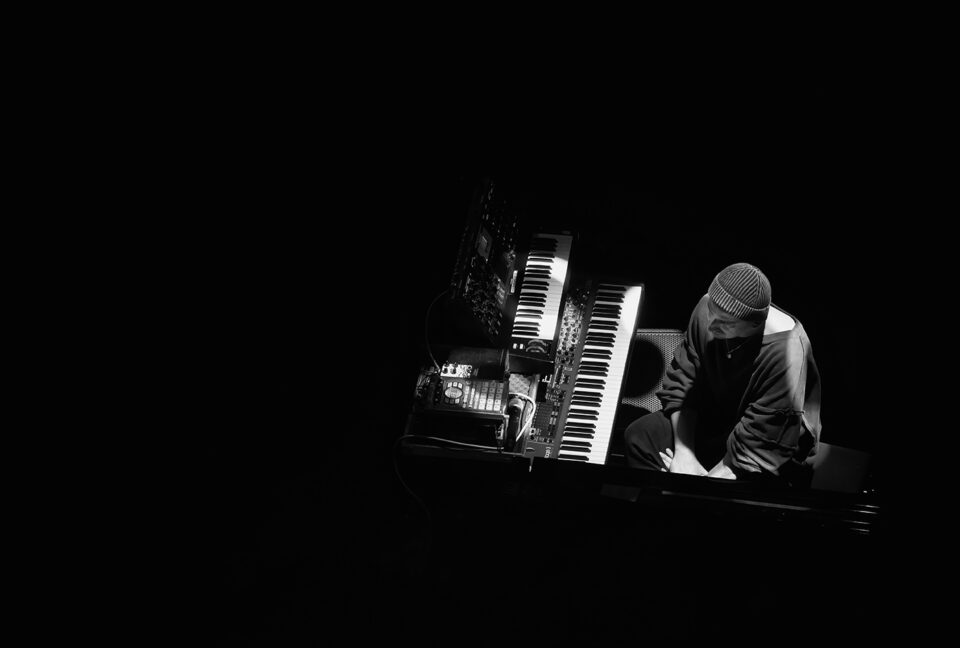 Czarno-białe, artystyczne zdjęcie muzyka w ujęciu z góry. Mężczyzna jest w luźnej bluzie i czapce. Siedzi przy zestawie syntezatorów z dwiema klawiaturami podobnymi do fortepianowych. Mężczyzna i instrumenty są delikatnie oświetlone i wyłaniają się z zupełnie czarnego tła.