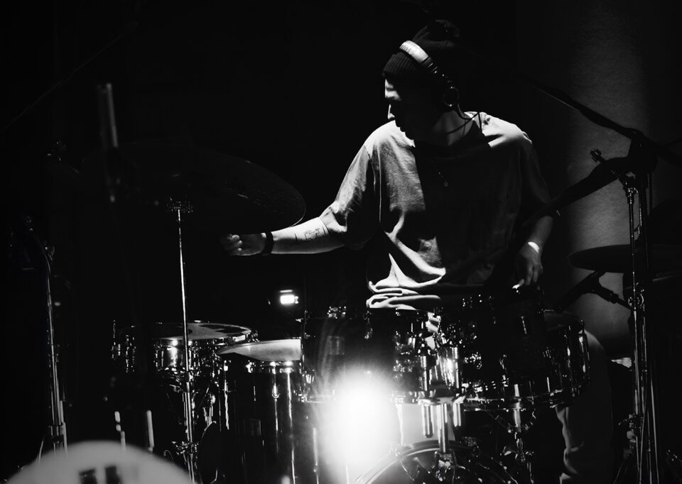 Czarno-białe zdjęcie perkusisty siedzącego za bębnami i talerzami. Perkusista w jednej ręce trzyma pałeczkę do gry, drugą ręką przytrzymuje jeden z talerzy na statywie. Ma luźną bluzę i czapkę. Tło ciemne.