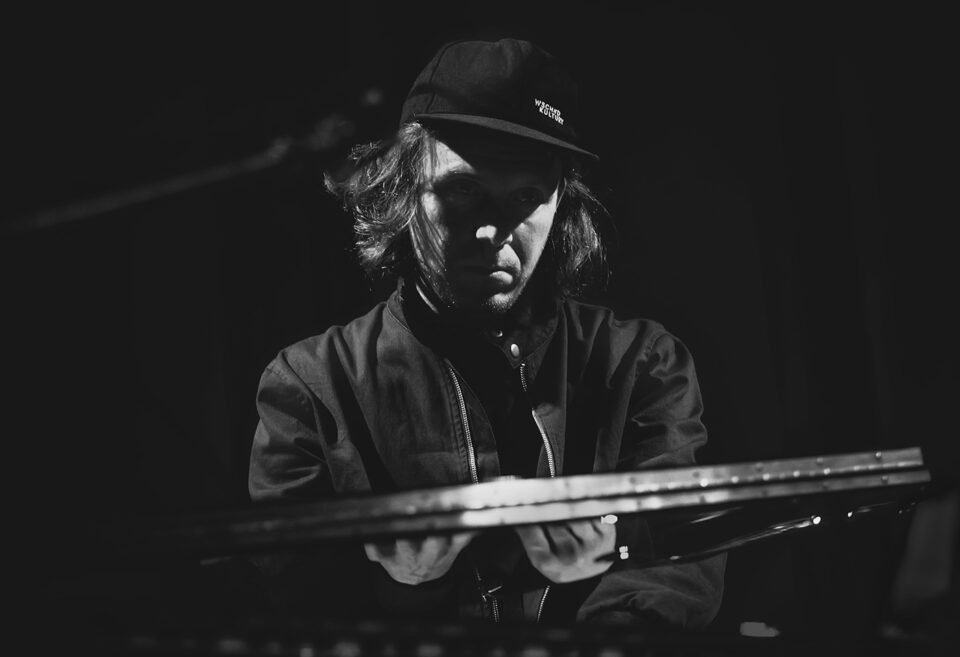 Czarno-białe zdjęcie portretowe muzyka. Przed nim znajduje się syntezator. Mężczyzna ma ciemną kurtkę, czapkę z daszkiem i jasne włosy sięgające za ucho. Tło Ciemne.