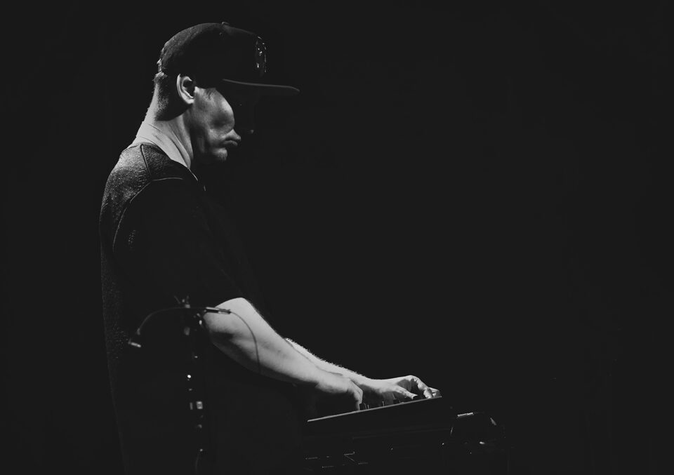 Czarno-białe zdjęcie muzyka w ujęciu od bioder w górę z profilu. Mężczyzna trzyma dłonie na syntezatorze. Ma koszulkę z krótkim rękawem i czapkę z daszkiem. Tło czarne.