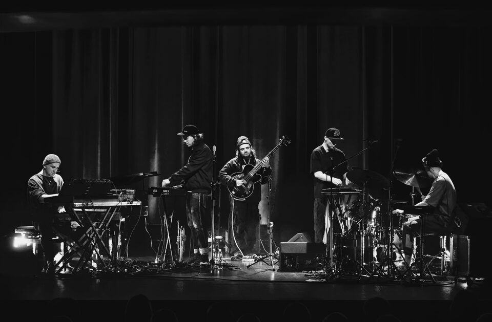 Czarno-białe zdjęcie pięcioosobowego zespołu na scenie. Klawiszowiec, trębacz, basista, saksofonista i perkusista. W tle ciemna kotara.