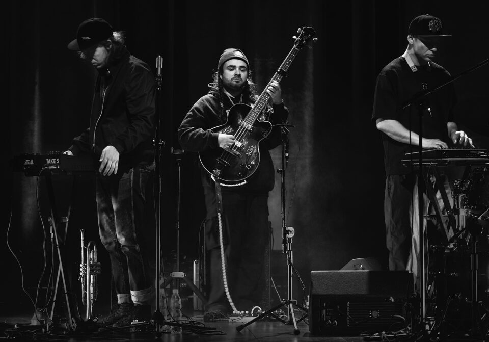 Czarno-białe zdjęcie trzech muzyków na scenie. W centrum basista, po bokach muzycy używający syntezatorów umieszczonych na statywach. Wszyscy są w ciemnych strojach i czapkach. Tło jest ciemne.