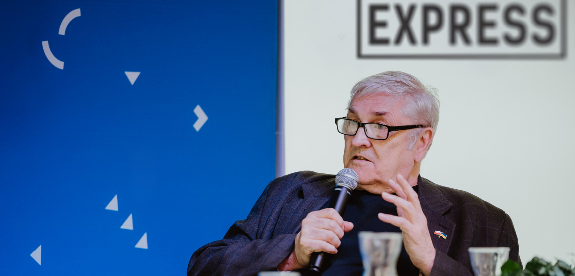 Zdjęcie siwowłosego mężczyzny w ciemnym stroju i okularach. Jest w ujęciu z półprofilu. Mężczyzna w jednej ręce trzyma mikrofon, drugą ręką gestykuluje. Tło podzielone na pół po lewej niebieskie, po prawej białe z czarnym napisem: 