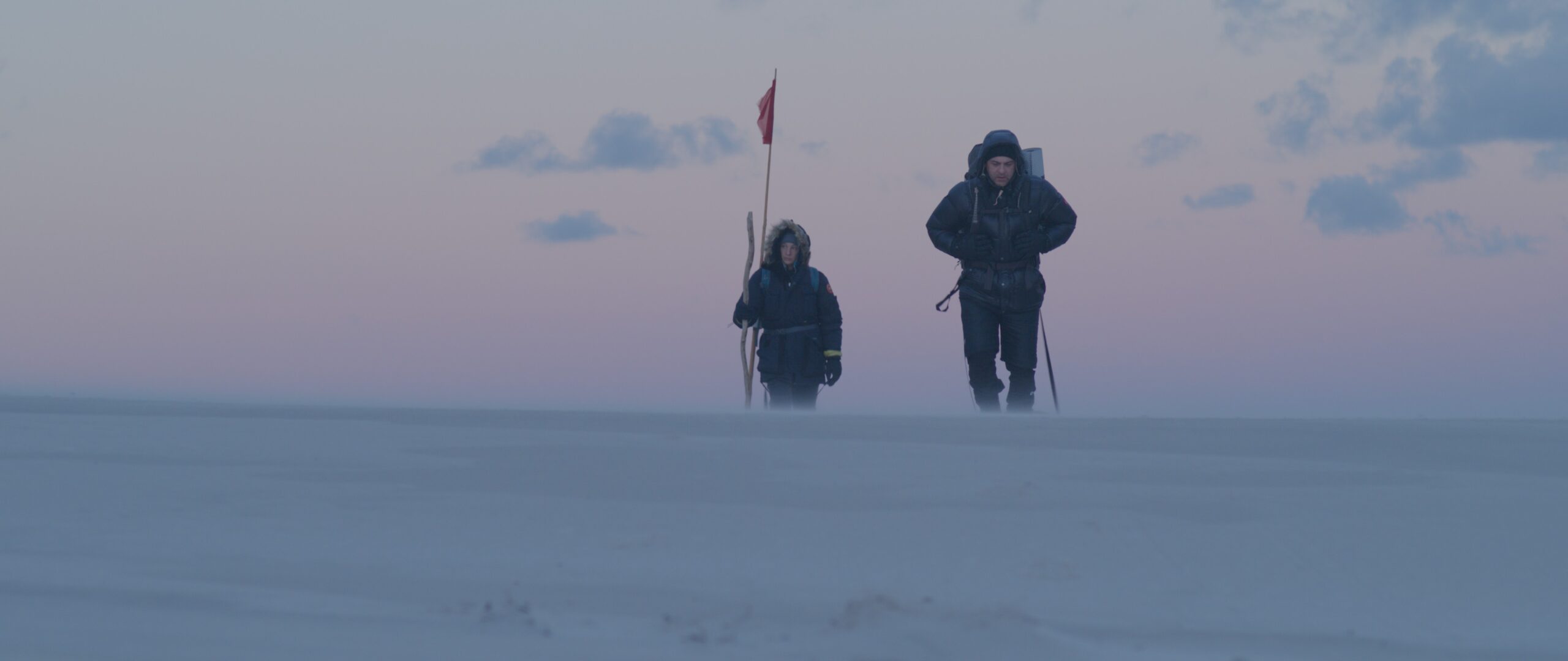 Zdjęcie dwóch osób w arktycznym krajobrazie. Postacie mają ciemne, grube stroje z kapturami i plecaki. Jedna z postaci trzyma czerwoną chorągiewkę na wysokim drzewcu. Wokół ośnieżone, równe podłoże. U góry niebo w ciepłym odcieniu.
