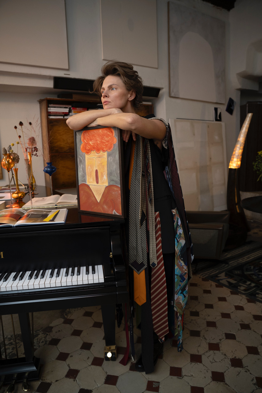 Zdjęcie kobiety w pomieszczeniu z obrazami na ścianach i starą podłogą z kamiennych płytek. Po lewej fortepian. Po prawej kobieta. Kobieta ustawiła obraz abstrakcyjny na klapie fortepianu, opiera ręce i podbródek na obrazie. Kobieta jest w ciemnym stroju obwieszonym krawatami w różnych kolorach i wzorach.