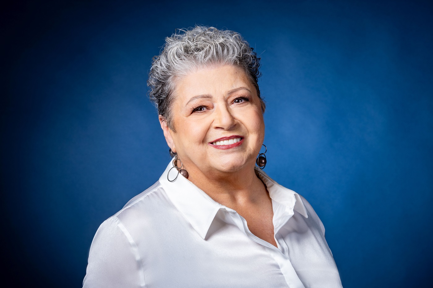 Zdjęcie portretowe uśmiechniętej kobiety. Ma krótkie, siwe włosy i duże kolczyki. Jest ubrana w białą koszulę z kołnierzem. Tło jest niebieskie.