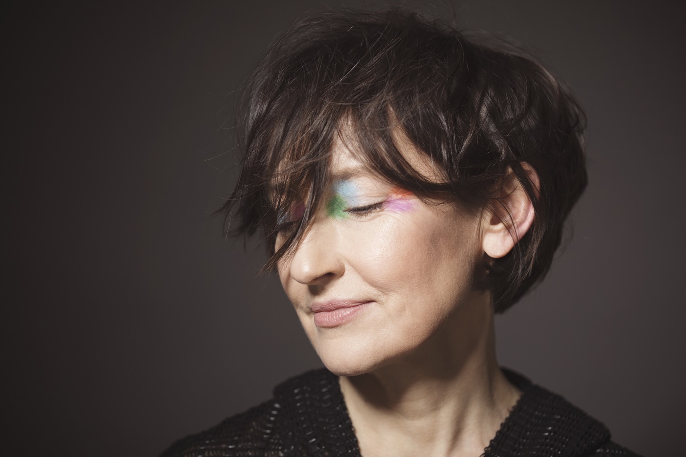 Zdjęcie portretowe uśmiechniętej kobiety w ujęciu z półprofilu. Kobieta ma ciemne włosy sięgające za ucho i czarny strój. Ma zamknięte oczy i kolorowe cienie na powiekach. Tło ciemnoszare.
