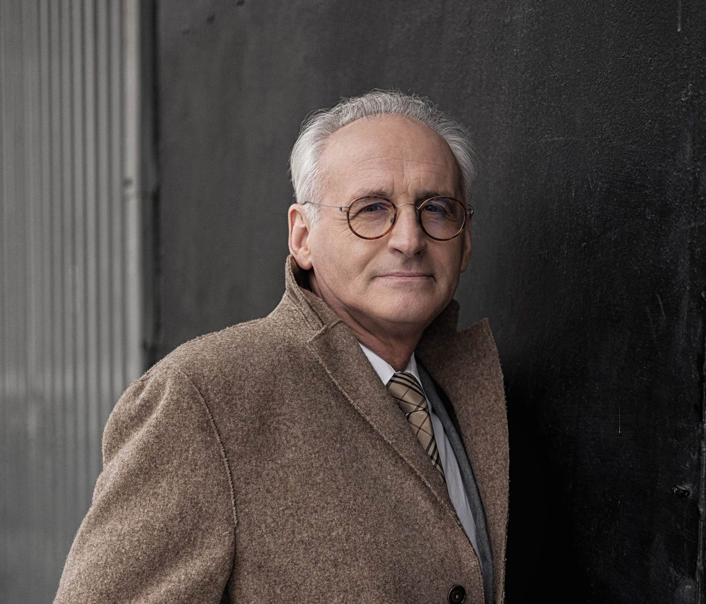 Zdjęcie portretowe mężczyzny. Ma siwe włosy, okulary i jest ubrany w białą koszulę z brązowym krawatem i brązową marynarką. Tło jest ciemnoszare.