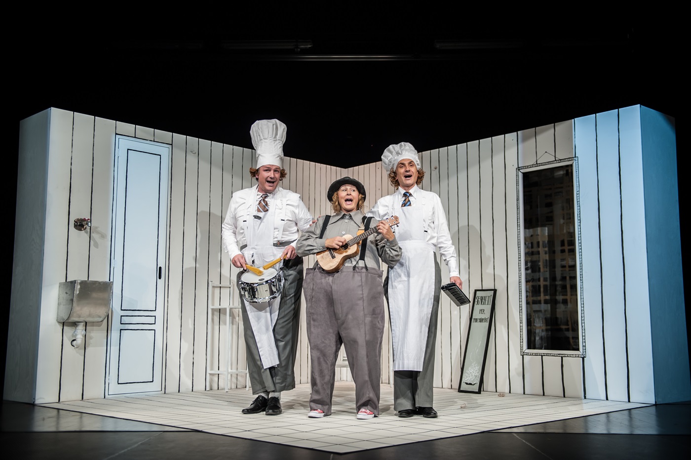 Zdjęcie trzech śpiewających osób. W centrum kobieta w szarym stroju ze spodniami na szelkach, gra na ukulele. Po bokach dwóch wysokich mężczyzn w białych strojach kucharzy z wysokimi czapkami. W tle scenografia z jasnym pomieszczeniem ze ścianami w pionowe pasy i drzwiami po lewej stronie.