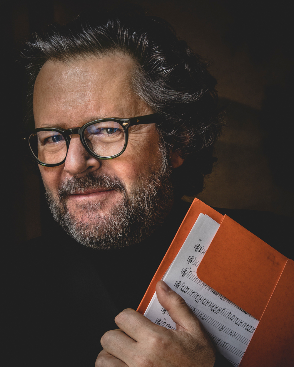 Zdjęcie portretowe uśmiechniętego mężczyzny. Jest w czarnym stroju. Ma ciemne, kręcone włosy, krótka brodę i wąsy. Ma okulary w czarnych oprawkach. Jedno ramię zasłania trzymając przy nim nuty na czerwonej podkładce. Tło czarne.