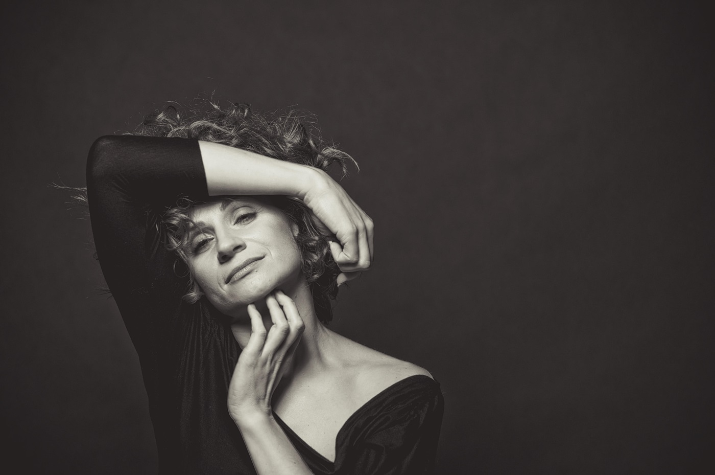 Czarno-białe zdjęcie kobiety w czarnym stroju. Kobieta ma kręcone włosy. Głowę ma lekko przechyloną w lewo. Jedną rękę trzyma nad czołem, drugą delikatnie dotyka podbródka. Tło jest czarne.