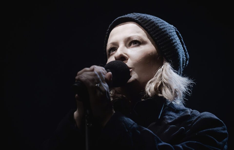 Zdjęcie kobiety. Zbliżenie na głowę i ramiona. Kobieta ma ciemne ubranie i czarną czapkę. Spod czapki wystają jasne włosy. Przed sobą trzyma czarny mikrofon. Tło czarne.