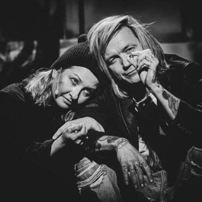 Obraz Główny: Czarno-białe zdjęcie kobiety i mężczyzny. Oboje przykucają obok siebie, kobieta opiera głowę na ramieniu mężczyzny. On trzyma przed sobą małą szklaną kulkę. Oboje na nią patrzą. Kobieta jest ubrana w ciemny płaszcz i czapkę, mężczyzna w podarte dżinsy i skórzaną kurtkę.