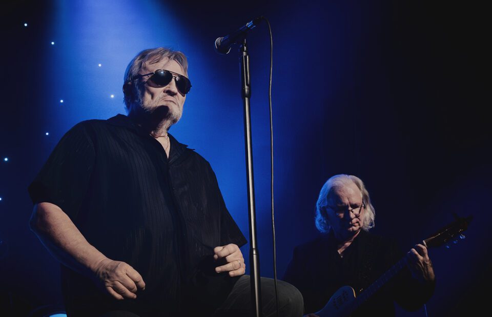 Dwóch muzyków podczas koncertu. Starsi mężczyźni w czarnych ubraniach. Wokalista w ciemnych okularach z mikrofonem na statywie oraz gitarzysta. Tło czarno-niebieskie.