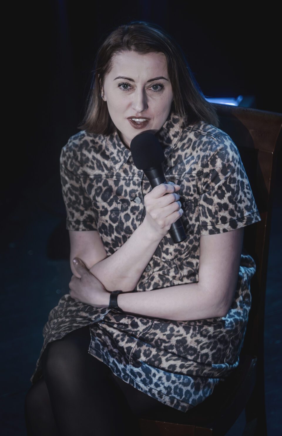 Zdjęcie kobiety siedzącej na krześle. Kobieta ma długie włosy. Jest ubrana w sukienkę z wzorem w panterkę z krótkim rękawem. Trzyma przed sobą mikrofon. Tło jest ciemne.