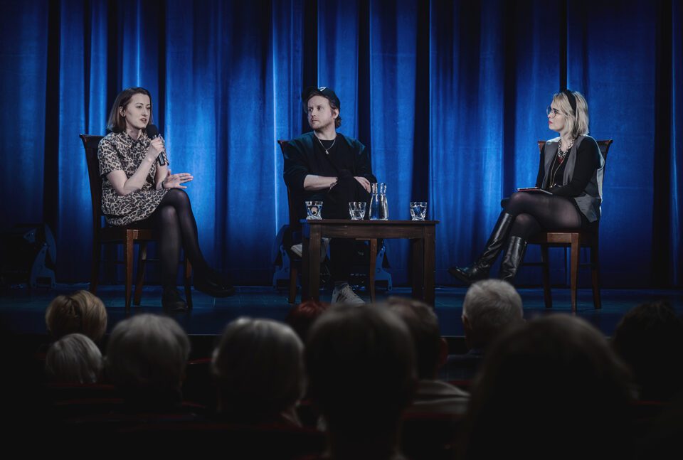 Ujęcie sceny zza pleców widzów. Na scenie trzy osoby siedzące na krzesłach. Po bokach dwie kobiety, w środku mężczyzna. przed mężczyzną stoi mały stolik. W tle kotara oświetlona na niebiesko.