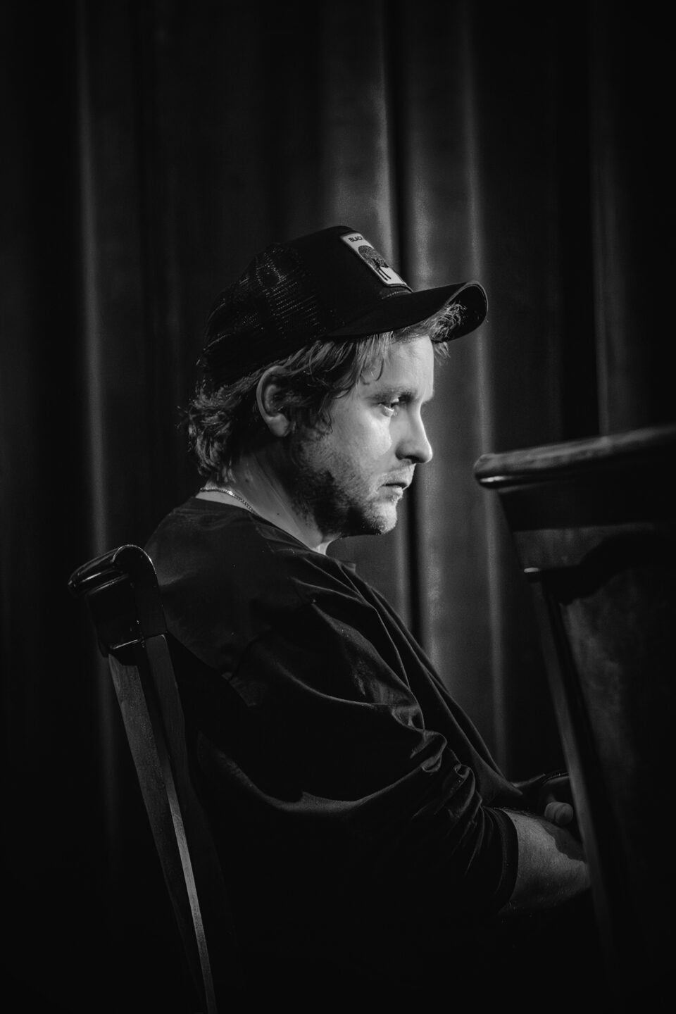 Czarno-białe zdjęcie młodego mężczyzny w ujęciu z profilu. Ma ciemną bluzę i czapkę z daszkiem. W tle ciemna kotara.