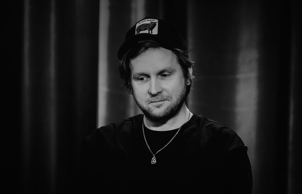 Czarno-białe zdjęcie portretowe młodego mężczyzny. Ma lekki zarost, jest ubrany w czarną bluzę i czapkę z daszkiem. W tle ciemna kotara.