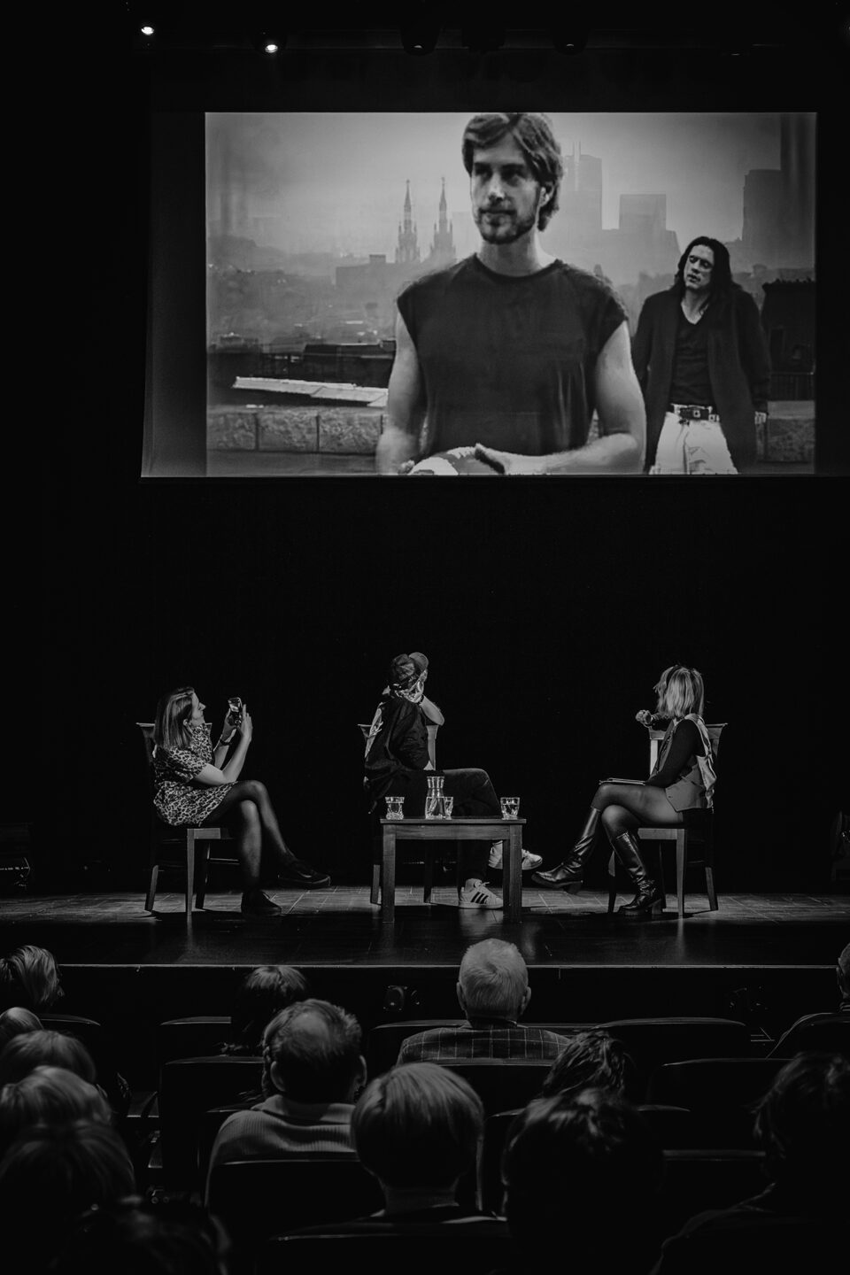 Zdjęcie czarno-białe. Ujęcie sceny zza pleców widzów. Na scenie trzy osoby siedzące na krzesłach. przed nimi po środku mały stolik. Nad nimi w głębi sceny duży ekran. Na ekranie dwóch mężczyzn z panoramą miasta w tle. Ekran wyłania się z czarnego tła.