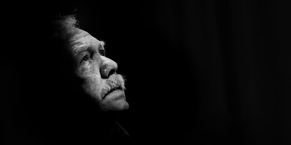 Slajd: Czarno-białe zdjęcie mężczyzny w ujęciu z profilu. Oświetlona jest tylko twarz kontrastująca z zupełną ciemnością w tle. Jest to straszy mężczyzna z jasnymi włosami i wąsami.