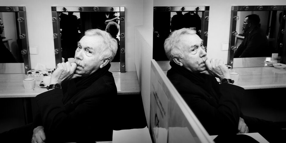 Slajd: Mężczyzna siedzący przy blacie w garderobie. Jest odwrócony plecami do ściany z dużym lustrem, w którym się odbija. Mężczyzna