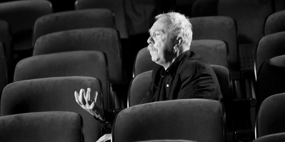 Slajd: Czarno-białe zdjęcie mężczyzny siedzącego na jednym z wielu foteli na widowni teatralnej. Mężczyzna ma jasne włosy i wąsy, jest ubrany na czarno. Odwraca głowę w lewo i gestykuluje. Fotele są ułożone w rzędy i zajmują całe tło zdjęcia.