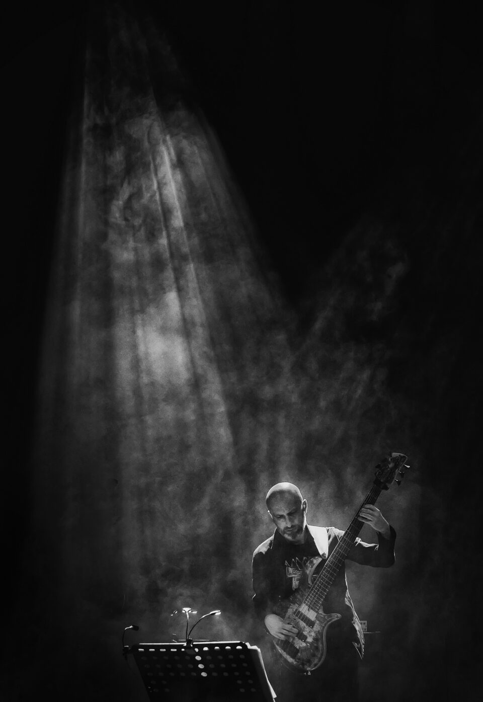 Czarno-białe zdjęcie mężczyzny grającego na gitarze basowej. Z góry na muzyka padają promienie delikatnego światła. Tło ciemne.