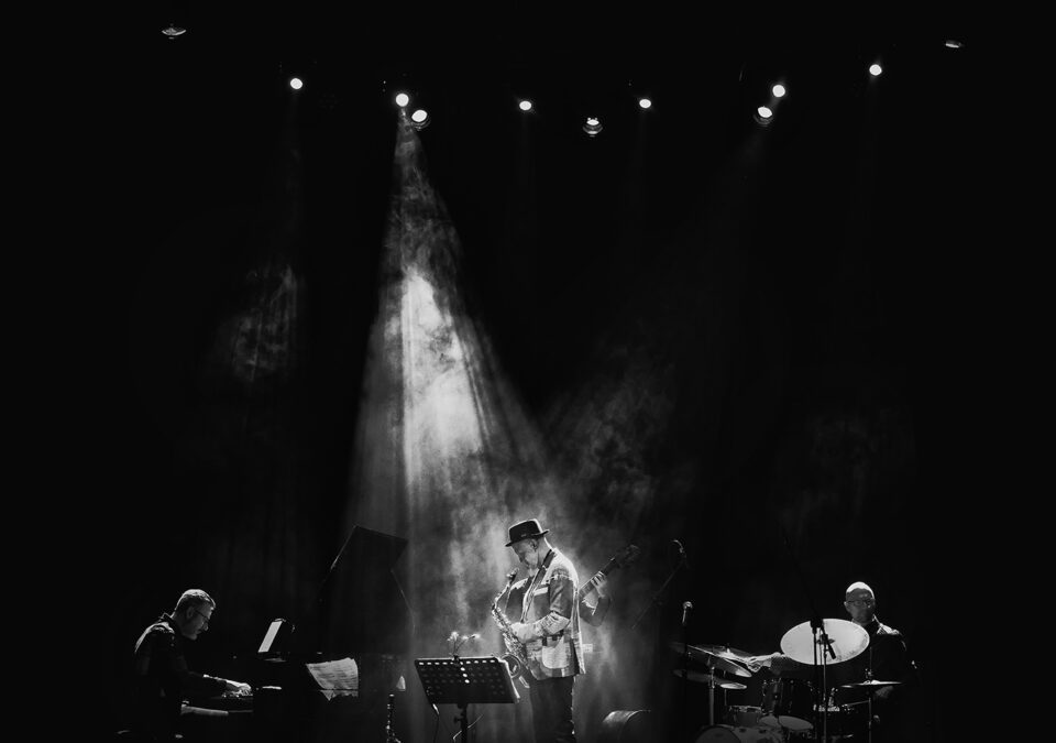 Czarno białe zdjęcie zespołu muzycznego na scenie. Po lewej pianista. Po środku saksofonista w strumieniu światła rozproszonego w sztucznym dymie. Zza saksofonisty wystaje ręka basisty i gryf basu. Po prawej saksofonista. Tło ciemne.