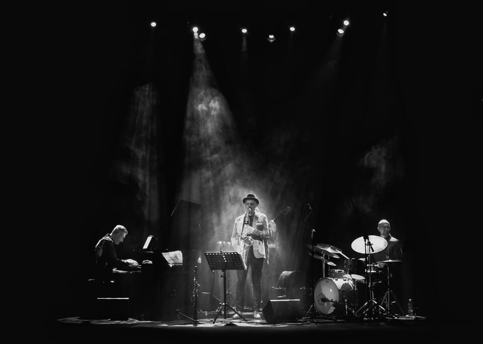 Czarno białe zdjęcie zespołu muzycznego na scenie. Po lewej pianista. Po środku saksofonista w strumieniu światła rozproszonego w sztucznym dymie. Po prawej saksofonista. Tło ciemne.