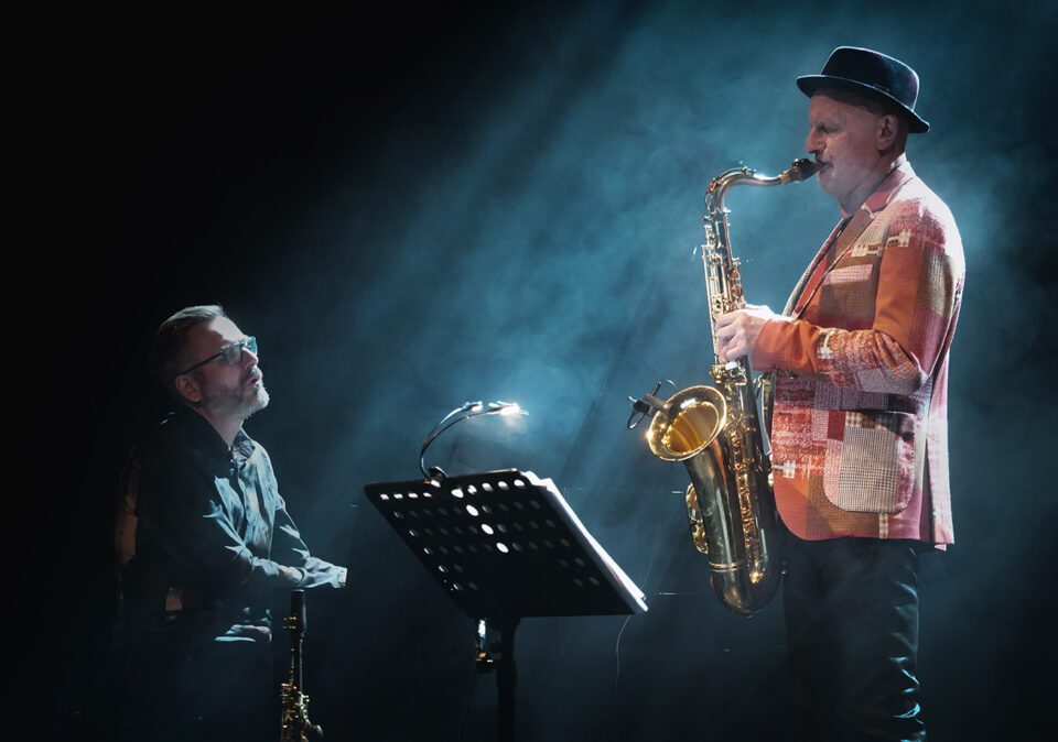 Zdjęcie dwóch muzyków zwróconych do siebie nawzajem. Po lewej pianista siedzący przy fortepianie. Po prawej saksofonista w kapeluszu. Tło ciemne z refleksami niebieskawego światła rozproszonymi w sztucznym dymie.