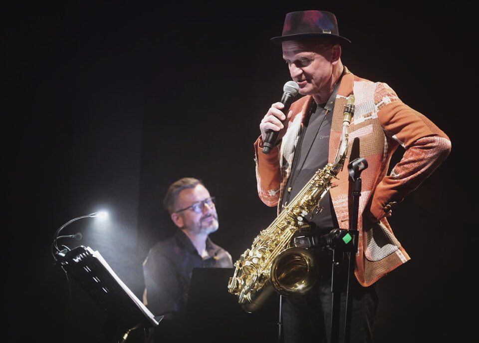 Zdjęcie dwóch muzyków. Na pierwszym planie mężczyzna z saksofonem zawieszonym na pasku przełożonym za szyją. Mężczyzna jedną ręką trzyma mikrofon, drugą podpiera bok. Ma pomarańczowo-beżową marynarkę i ciemny kapelusz. Na drugim planie po lewej pianista. Tło jest czarne.