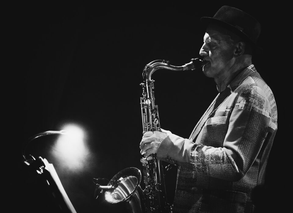 Czarno-białe zdjęcie grającego saksofonisty w ujęciu z profilu. Mężczyzna ma wzorzystą marynarkę i ciemny kapelusz. Tło czarne z rozbłyskiem światła w lewym dolnym rogu.