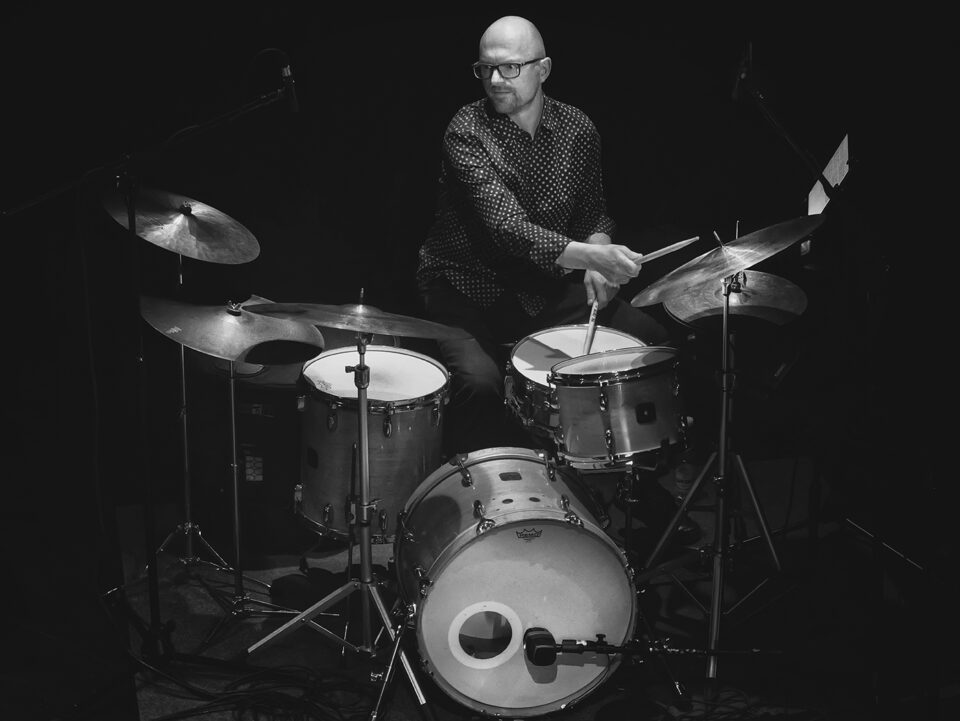 Czarno-białe zdjęcie perkusisty. Mężczyzna jest łysy, ma okulary. Jest ubrany w ciemne spodnie i ciemną koszulę w drobny deseń. Siedzi za zestawem